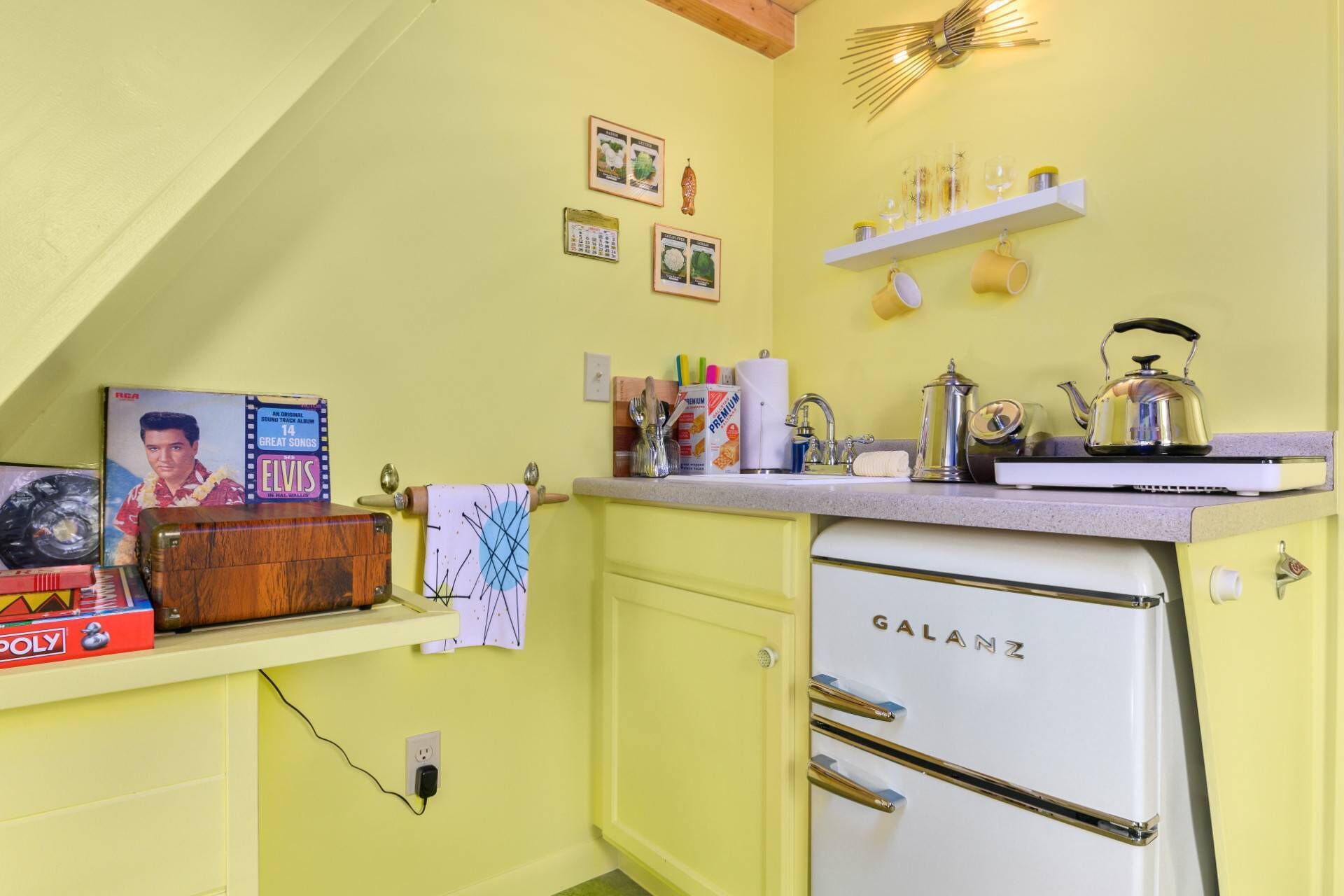 Uma cozinha compacta com geladeira Galanz, uma pia pequena e bancada com diversos itens, incluindo chaleira e cafeteira.