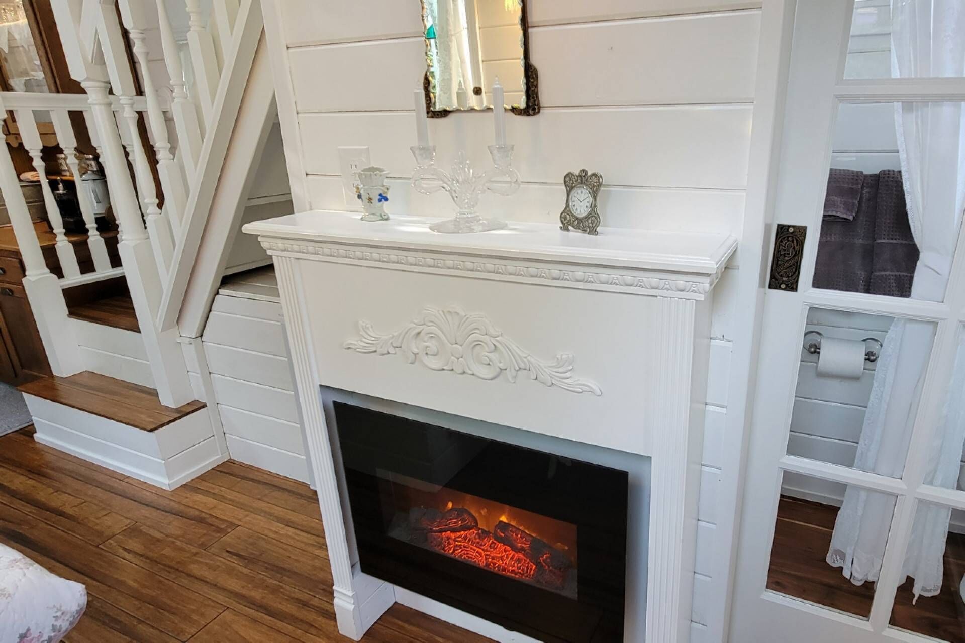 Una chimenea blanca con puerta de vidrio, un rodapié con adornos y una escalera con barandillas blancas.