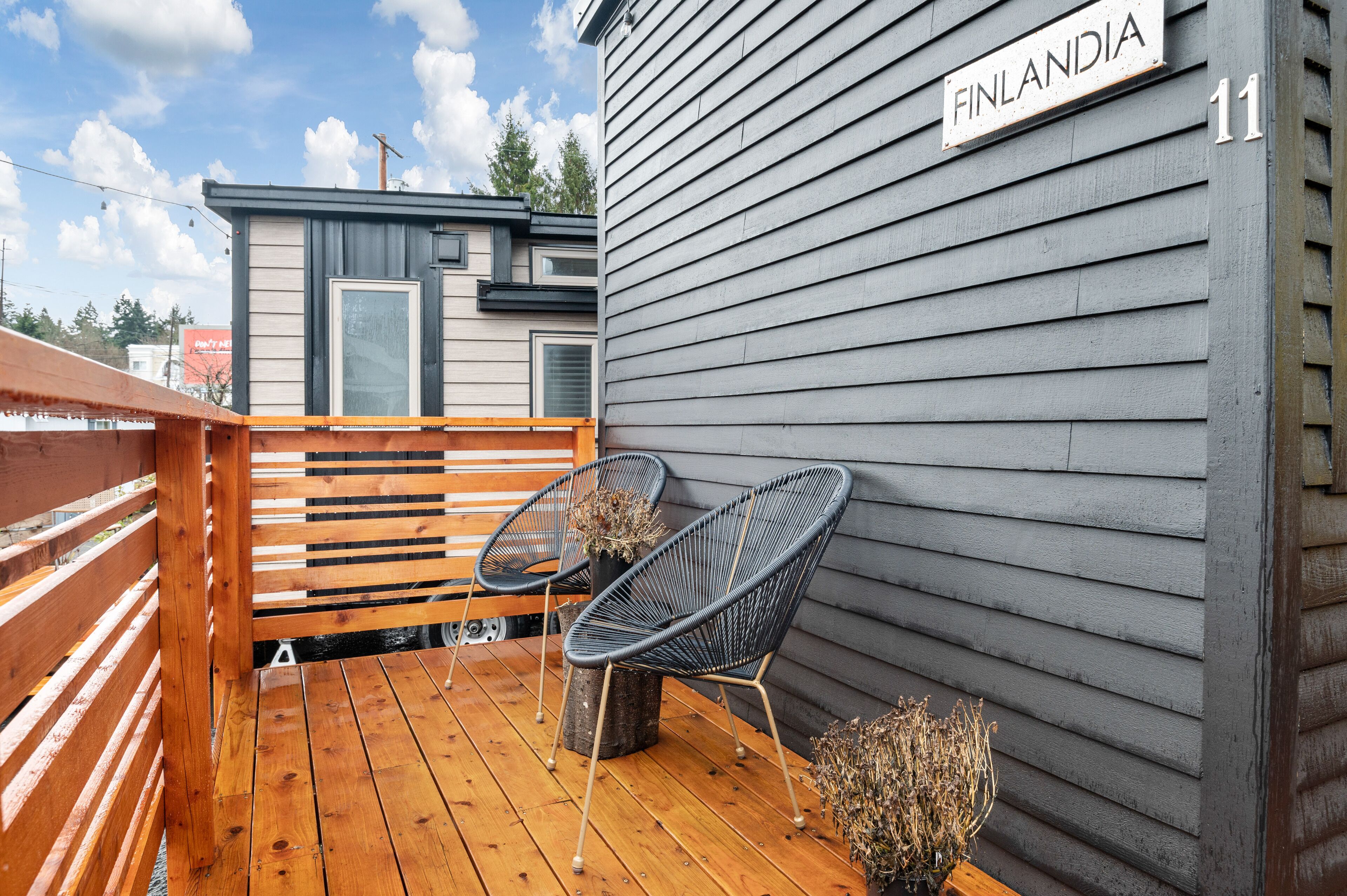 Una terraza de madera con dos sillas negras de alambre y una mesita, una casa gris con un letrero que dice 'FINLANDIA' y el número '11' en la pared.