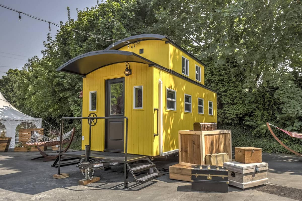 A yellow, two-story house with a curved roof, a front door, and windows. There are wooden crates and a hammock in front.