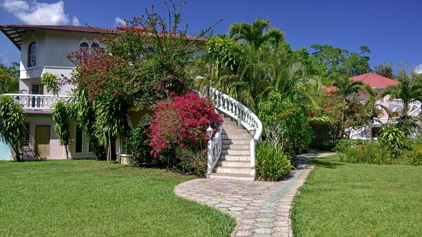 Una casa con una escalera curva que da a un balcón, rodeada de abundante vegetación y plantas en flor.
