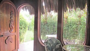 Una habitación con puerta de madera, una silla de mimbre, una mesa de vidrio y vista a un jardín a través de una ventana.