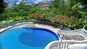 Una gran piscina azul clara rodeada por una balaustrada blanca, con abundante vegetación y un edificio al fondo.