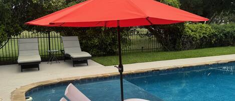 A pool area with a red umbrella, two lounge chairs, and a poolside setting.