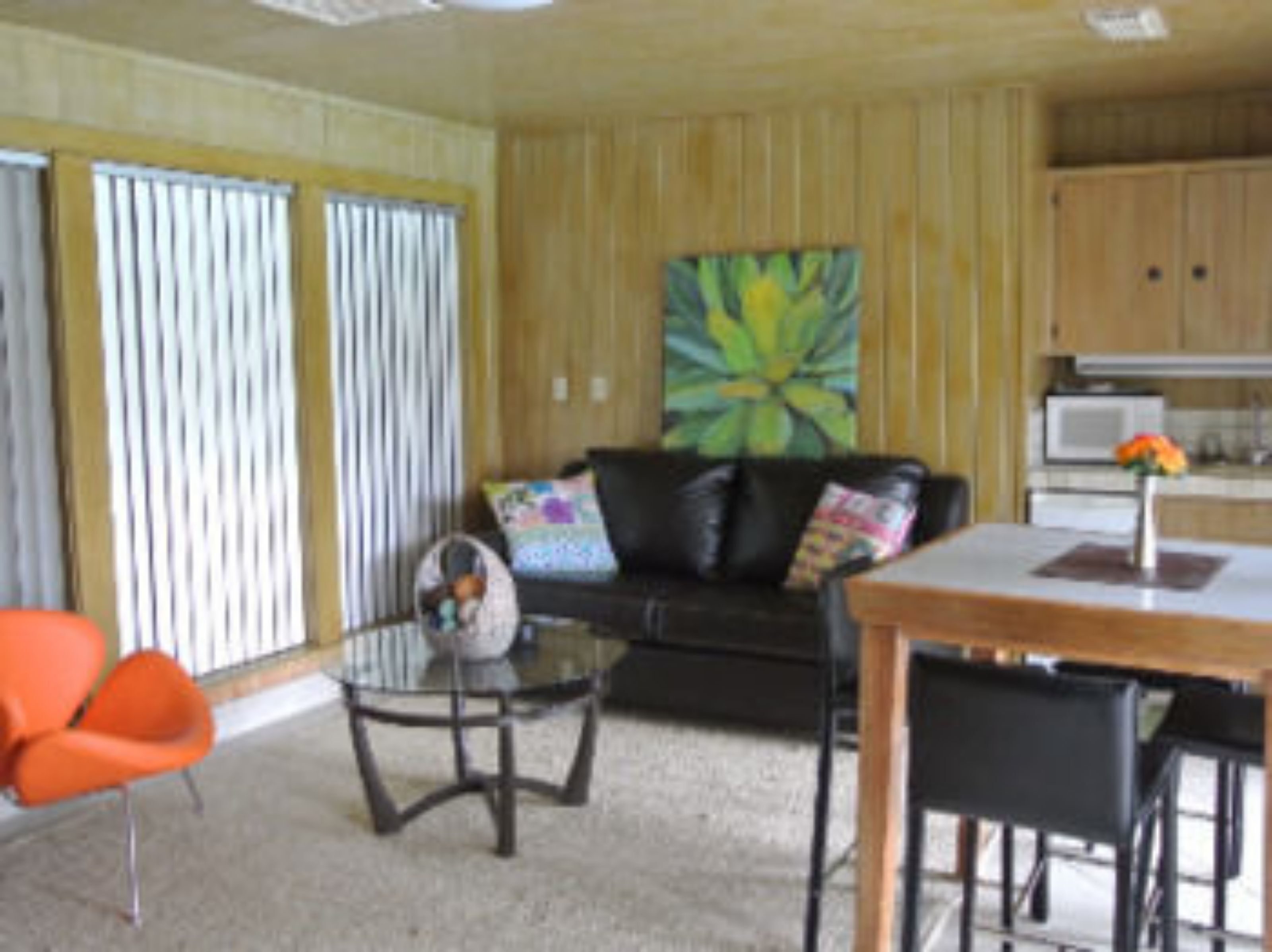 A living room with a black leather sofa, a glass coffee table, and orange chairs. There is a kitchen area with wooden cabinets and a microwave.