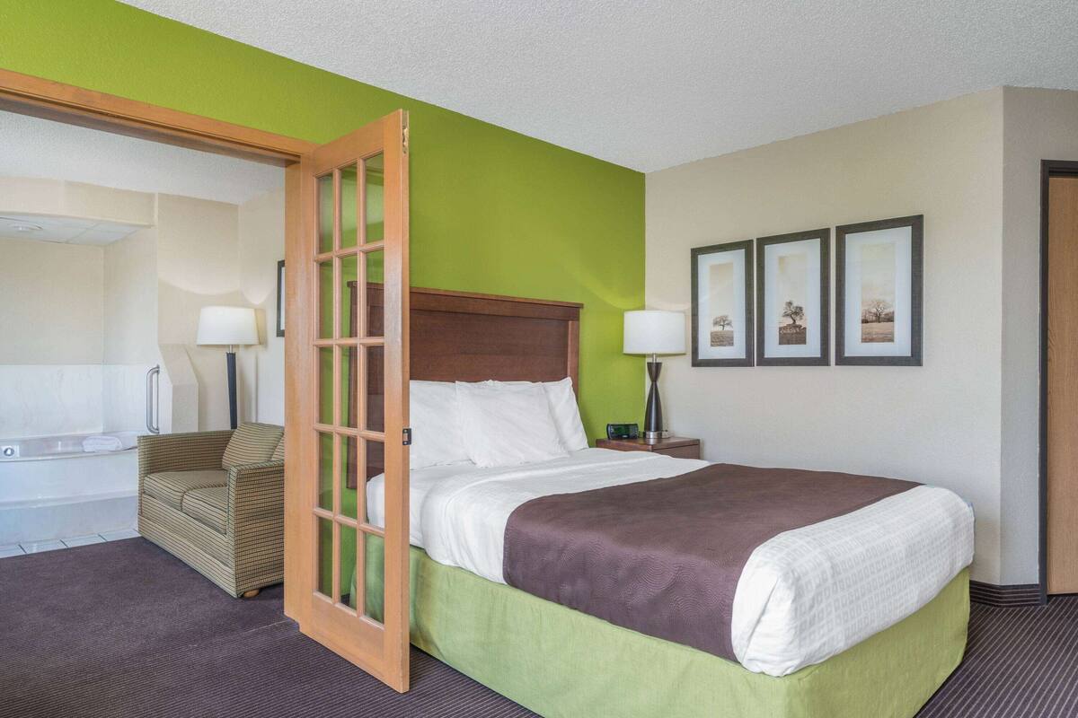 A hotel room with a bed, a green wall, a wooden headboard, a green sofa, a small table with a lamp, and a framed picture on the wall.