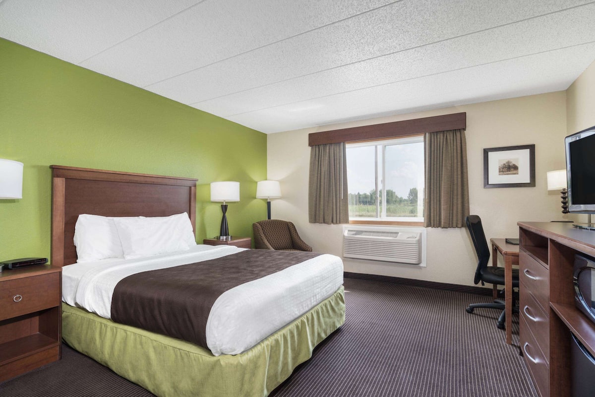 A hotel room with a large bed, two bedside tables with lamps, a desk with a chair, a television, and a window with curtains.