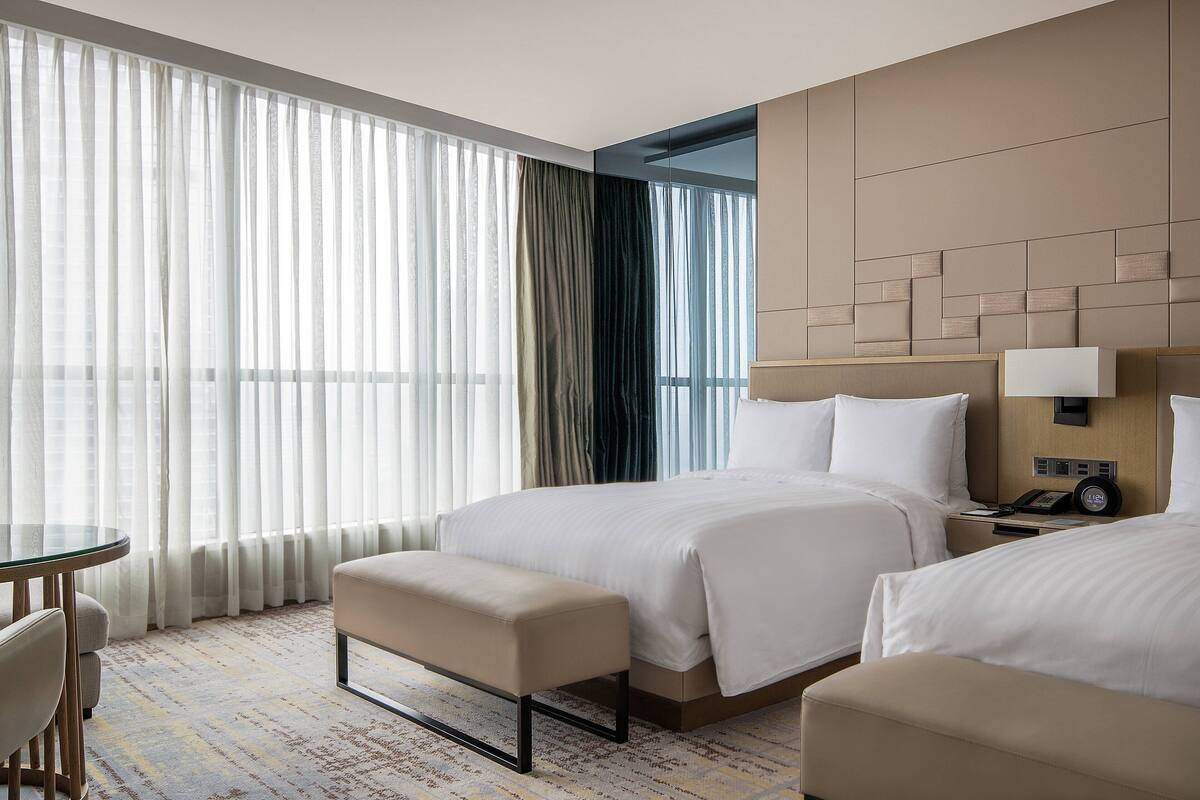 Ein Hotelzimmer mit zwei Betten, einem Nachttisch mit Lampe, einem kleinen Tisch mit Stühlen und einem großen Fenster mit durchsichtigen Vorhängen.