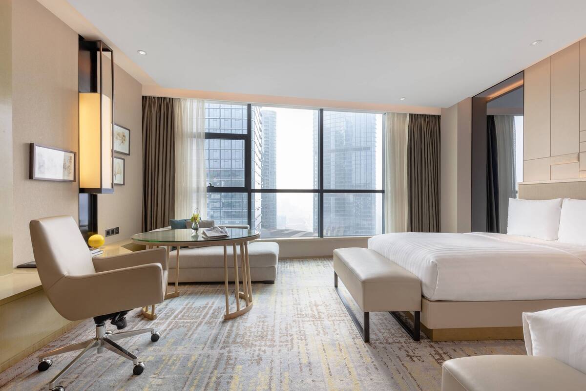 Ein Hotelzimmer mit einem großen Bett, einer Sitzecke, einem Schreibtisch und Blick auf hohe Gebäude durch große Fenster.
