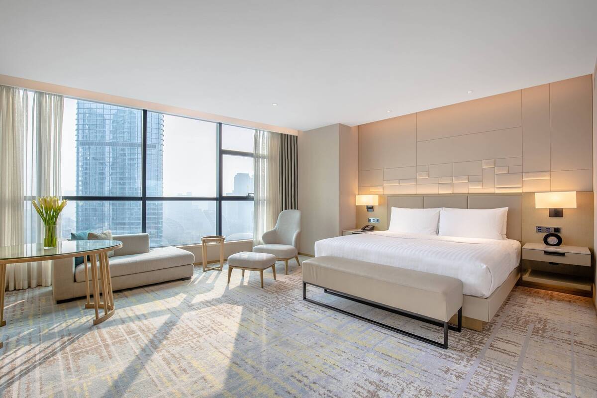 Ein Hotelzimmer mit einem großen Bett, einem Schlafsofa, einem Sessel, einem kleinen Tisch und Blick auf eine Stadtkulisse durch große Fenster.