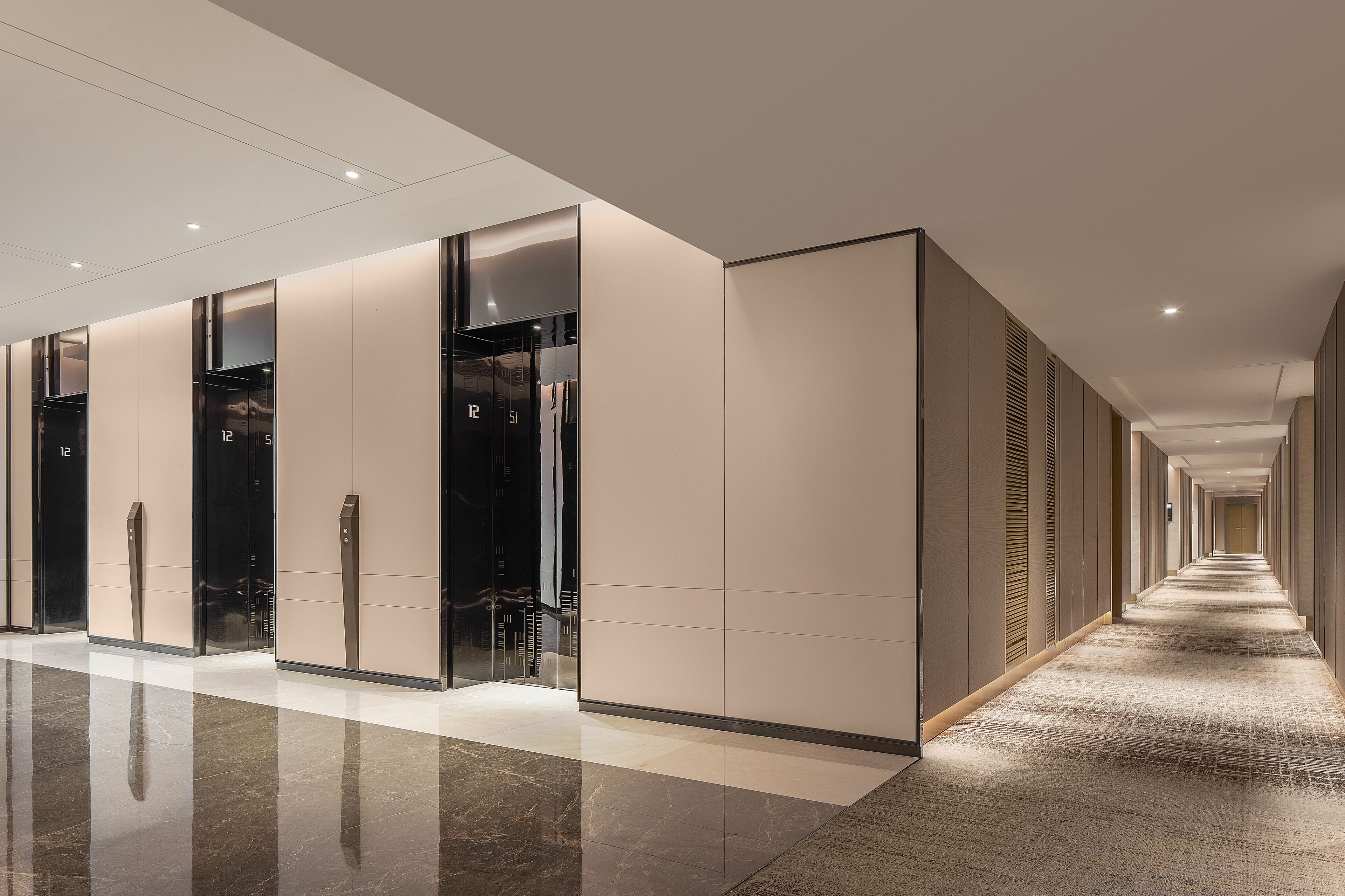 A modern hotel corridor with a long, straight layout, featuring a series of elevators and a carpeted floor.