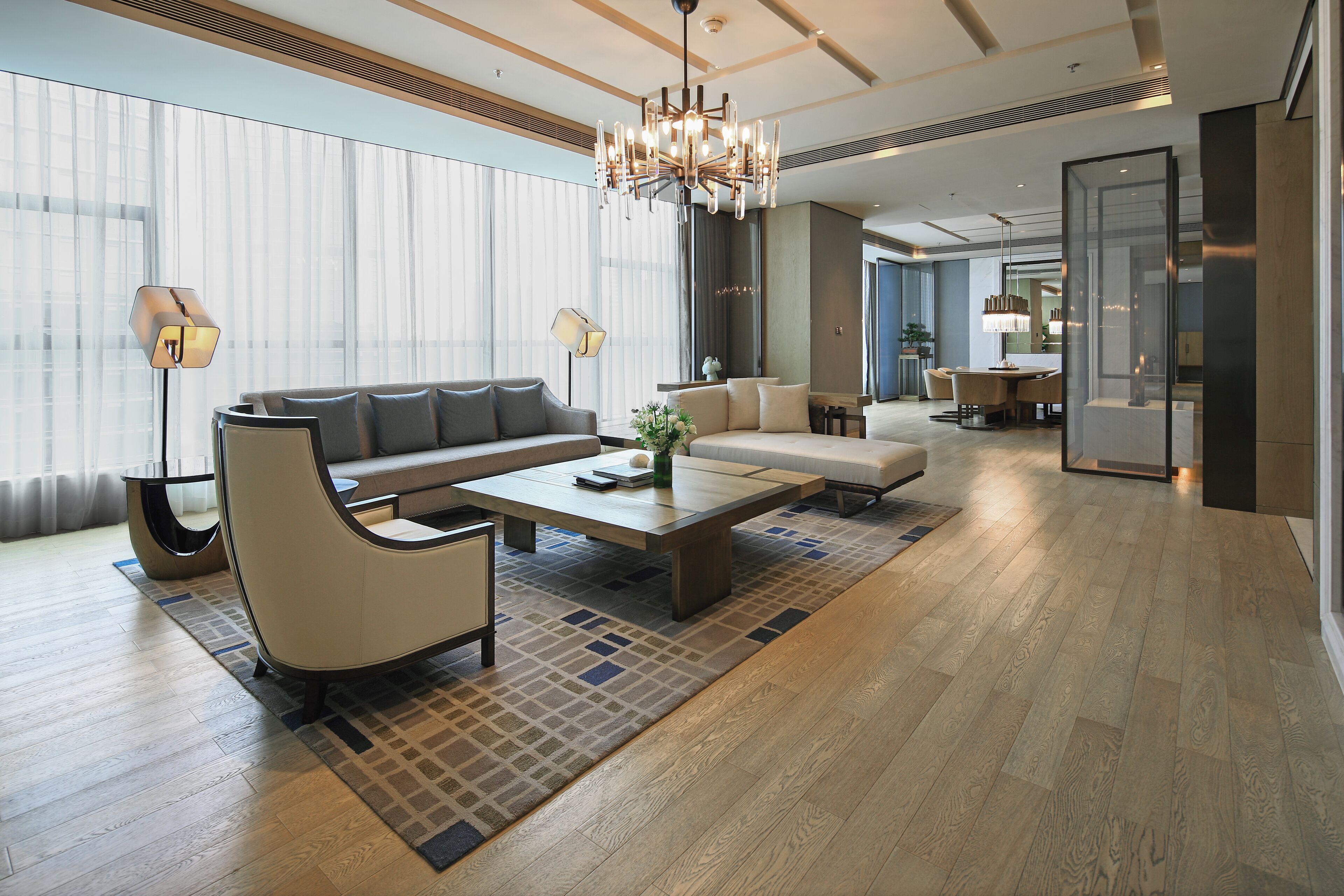 A modern hotel lobby with a large sofa, coffee table, and floor lamp. There is a dining area with a table and chairs in the background.