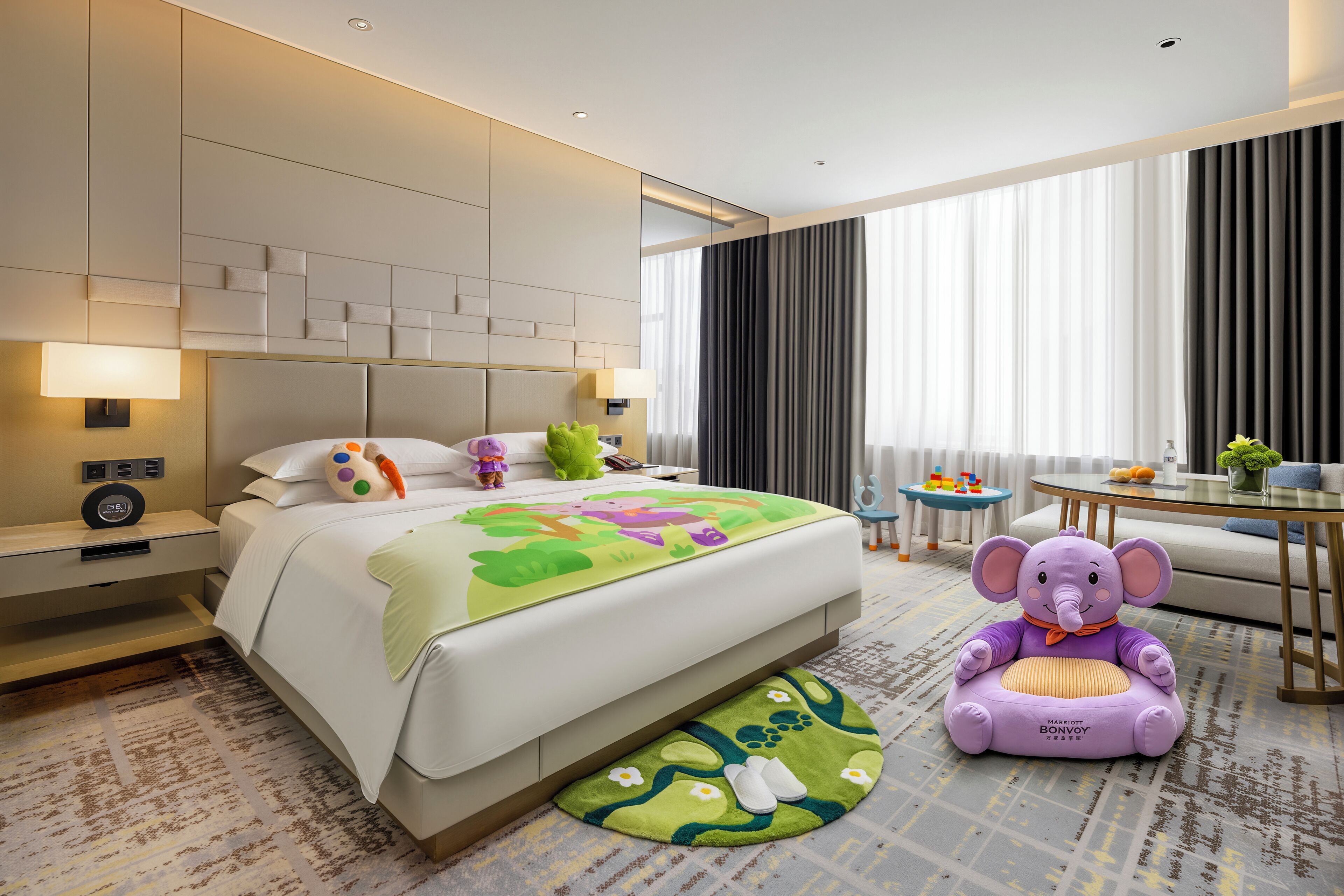 A hotel room with a bed, bedside tables, a nightstand, a sofa, a small round table, and a plush elephant toy.