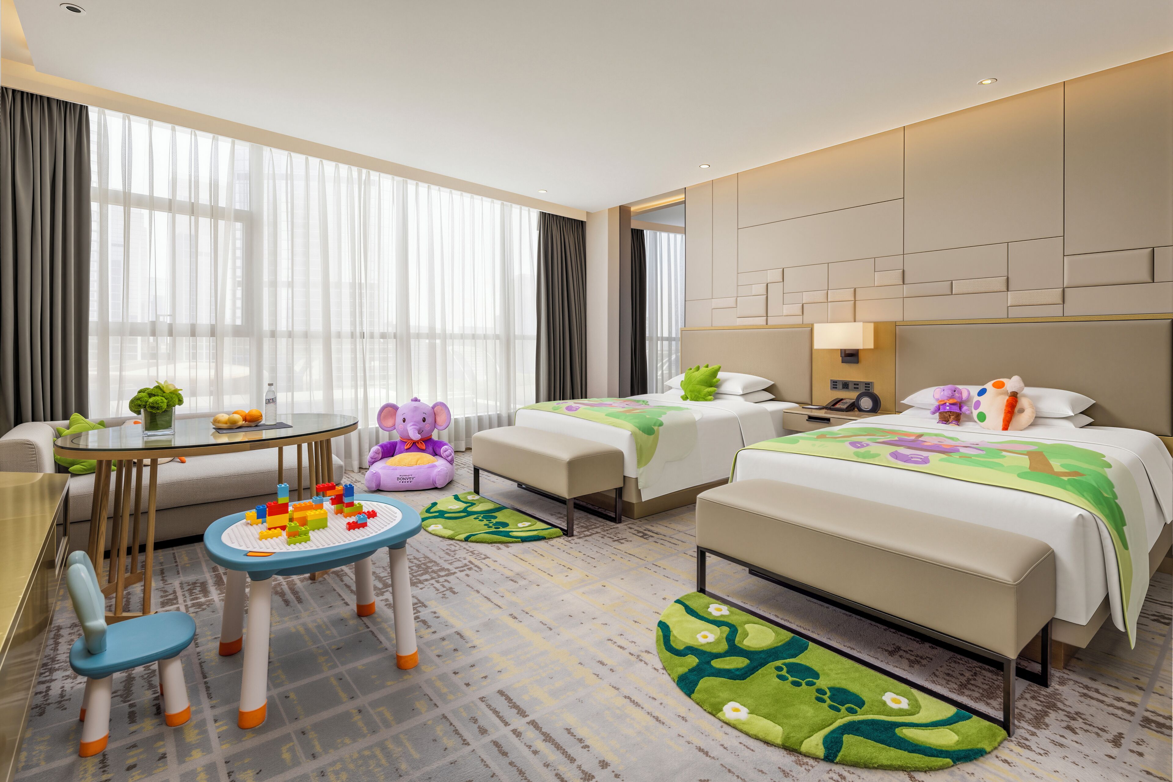 A hotel room with two beds, a small table, a chair, and a play area with toys.