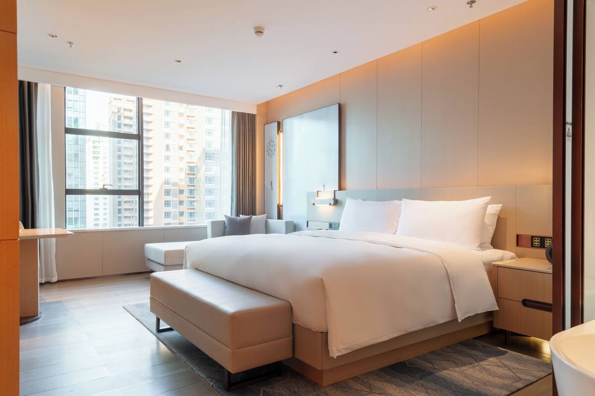 Ein modernes Hotelzimmer mit einem großen Bett, einem Nachttisch, einem Sessel und Blick auf hohe Gebäude durch das Fenster.