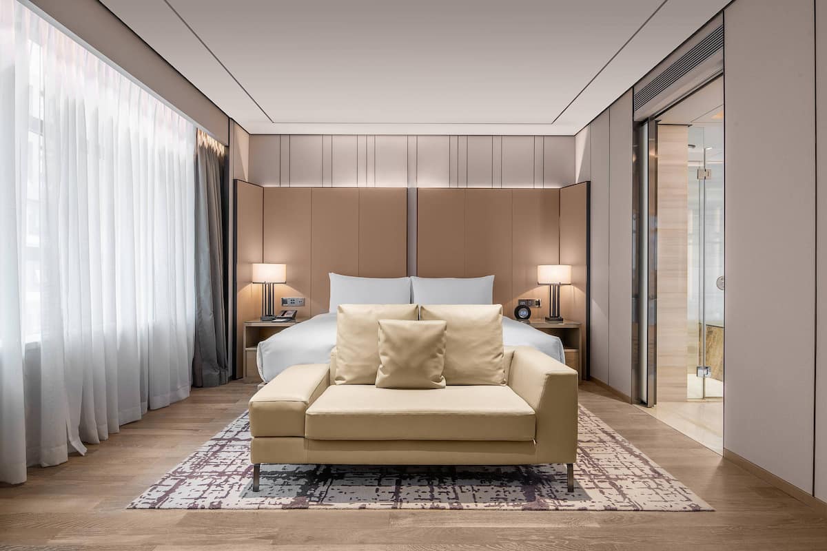 Ein modernes Hotelzimmer mit einem großen Bett, zwei Nachttischen mit Lampen, einem beigefarbenen Sofa und einem gemusterten Teppich.