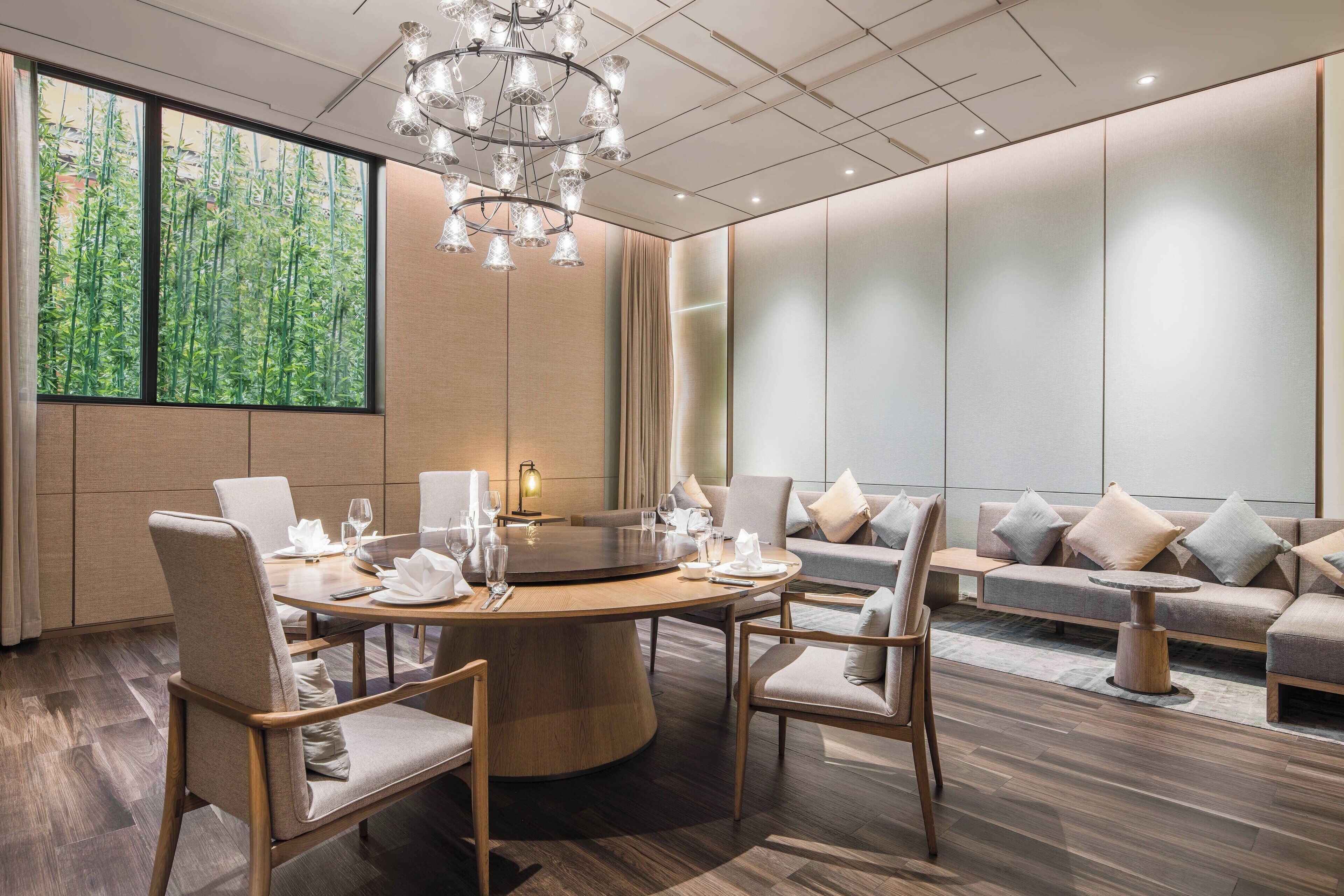A modern hotel room with a round dining table, comfortable seating, a large window with a view of bamboo, and a stylish chandelier.