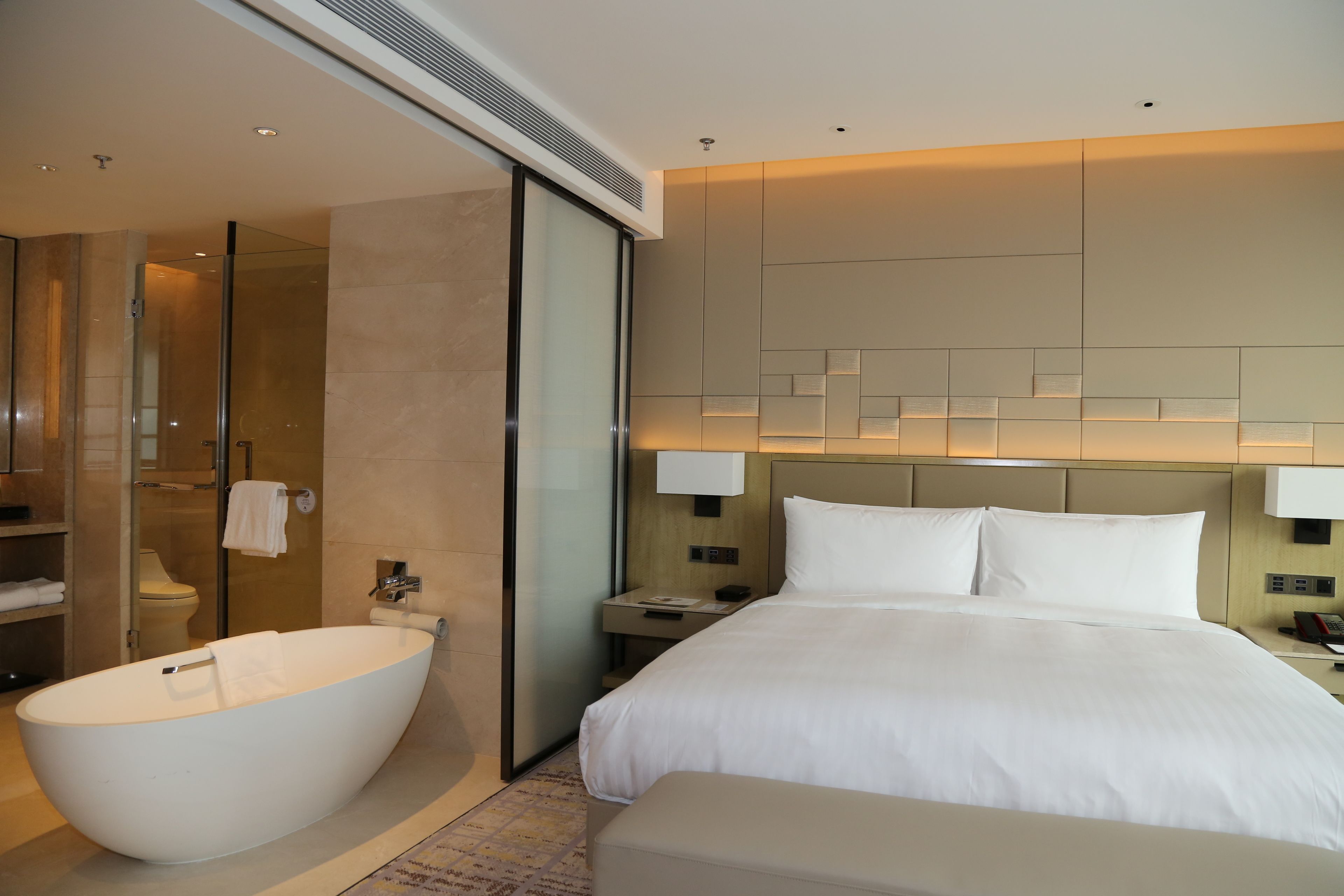 A modern hotel room with a large bathtub, a separate shower area, a neatly made bed, and a bedside table with a lamp.