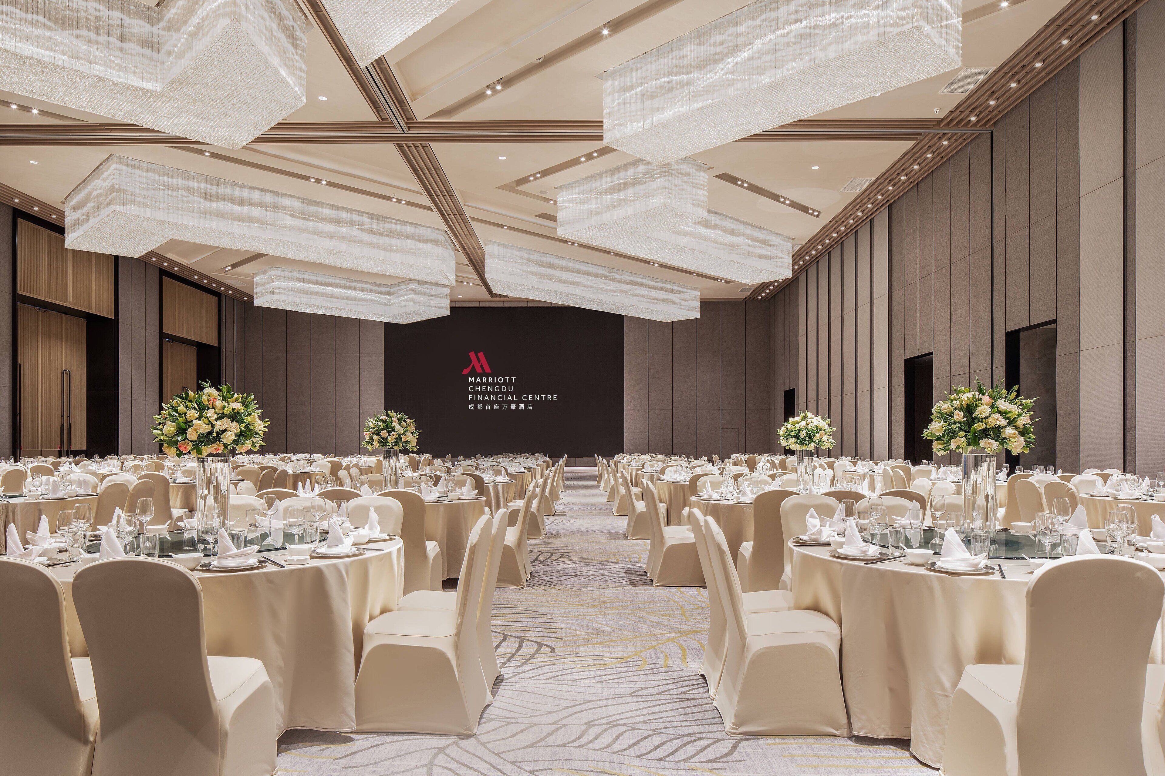 A banquet hall with round tables, beige chairs, and floral centerpieces.