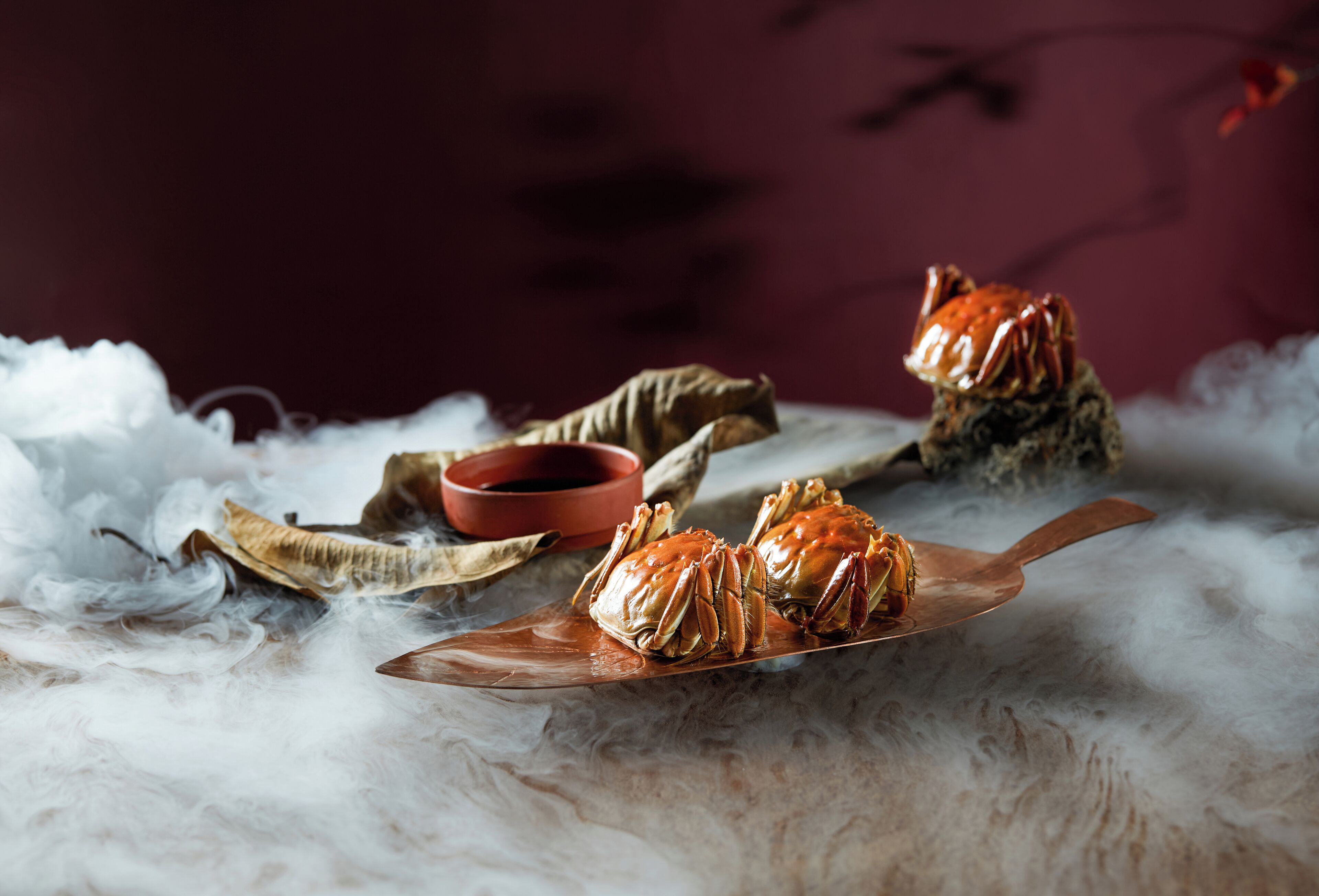 Two crabs on a copper plate with a red bowl and dried leaves in the background.