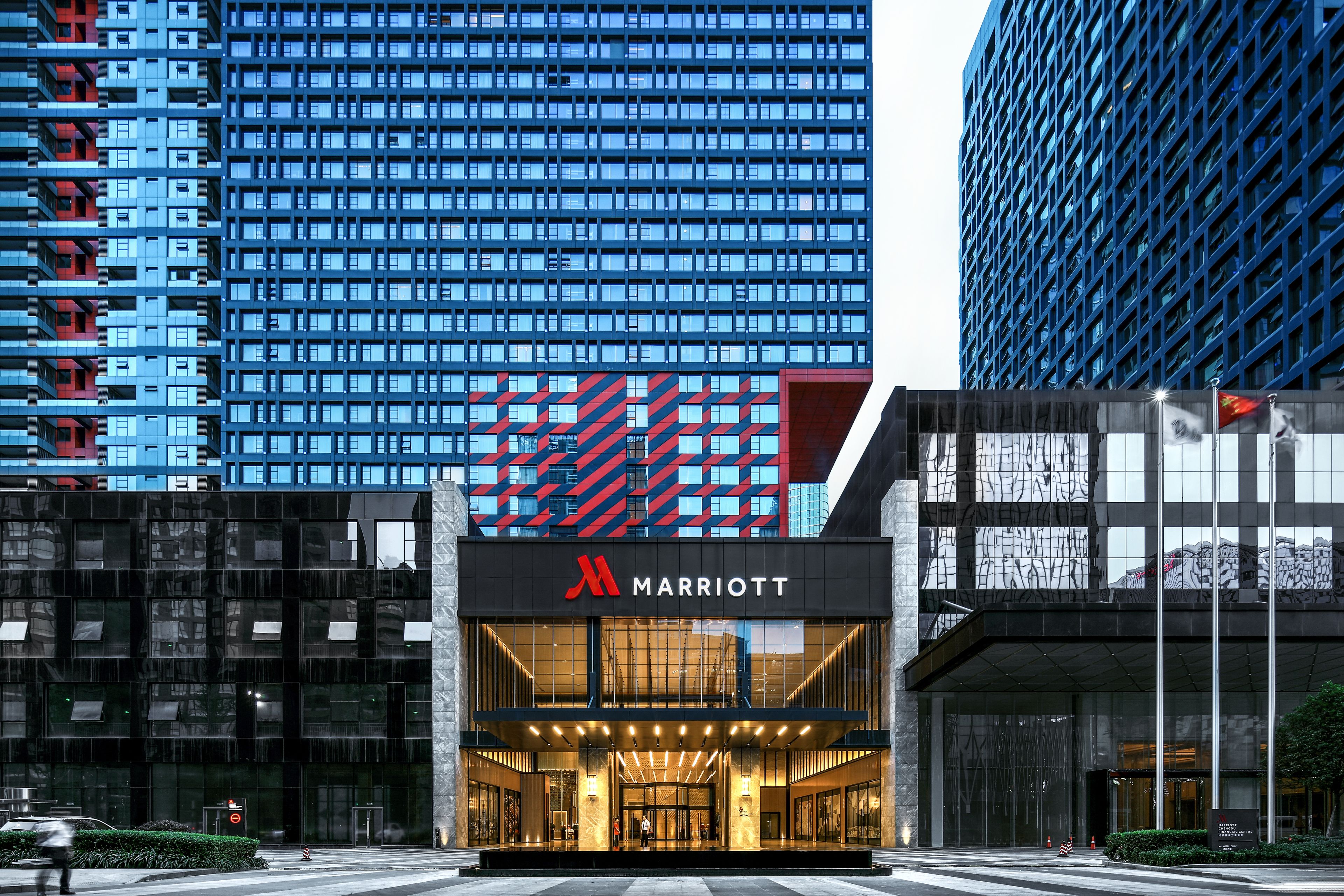 Der Eingang eines Marriott Hotels mit einer modernen Glasfassade, einem gut sichtbaren roten und blauen Logo und einer großen Glaseingangstür.