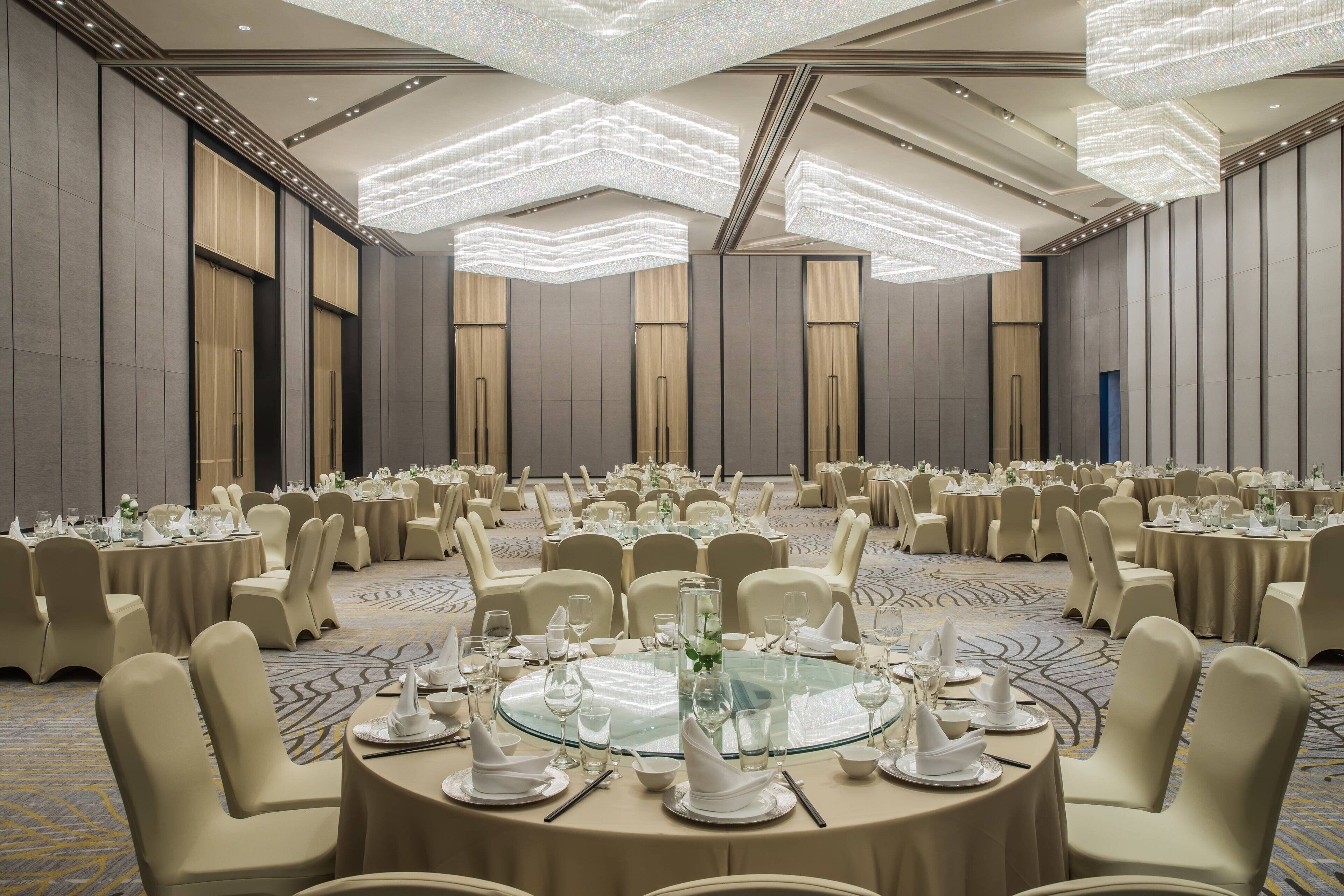 A large banquet hall with round tables set for dining, high ceilings with modern lighting fixtures, and a carpeted floor.