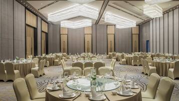 A large banquet hall with round tables set for dining, high ceilings with modern lighting fixtures, and a carpeted floor.