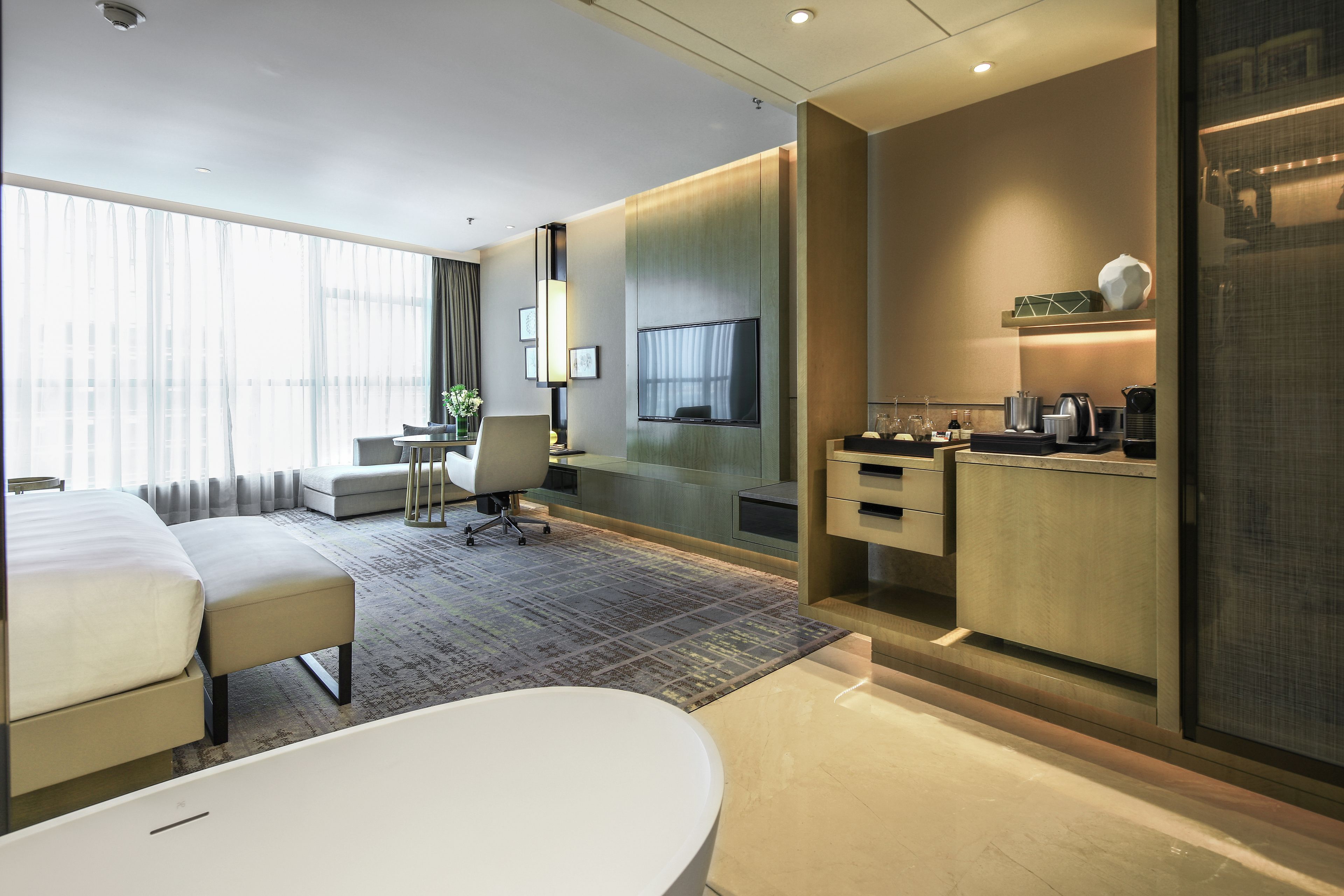 A modern hotel room with a large bed, a desk, a seating area, and a kitchenette.