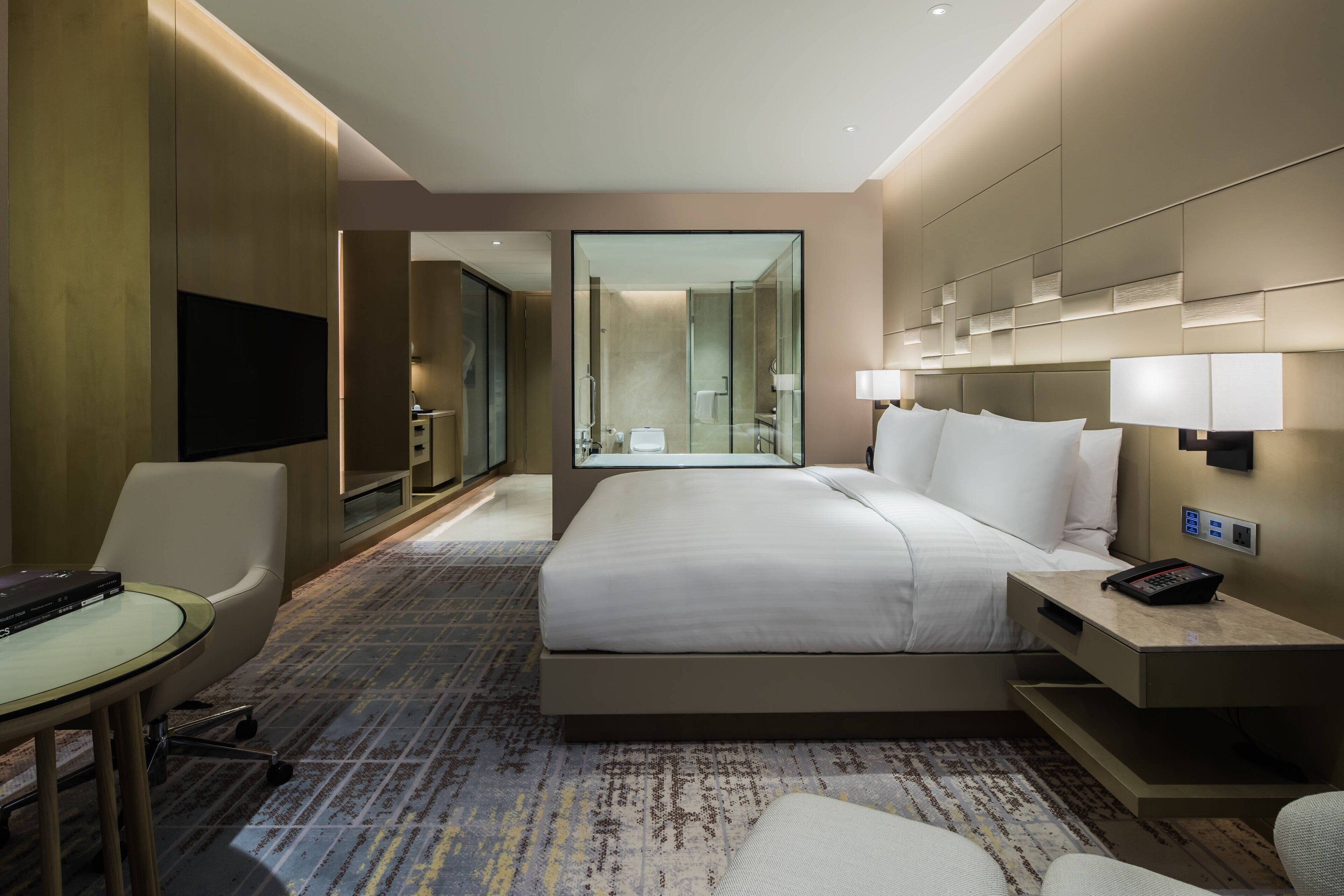 A modern hotel room with a large bed, a desk with a lamp, a chair, a TV, and a bathroom visible through a glass door.
