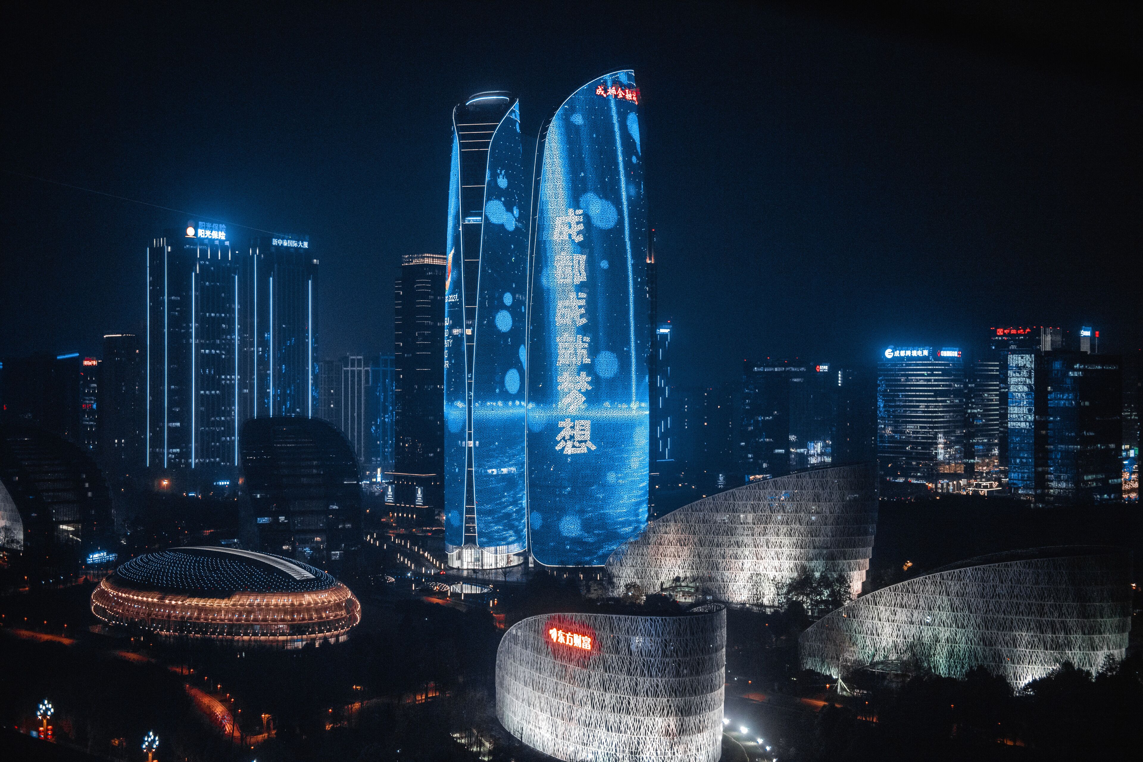 A cityscape at night with illuminated skyscrapers and a stadium.