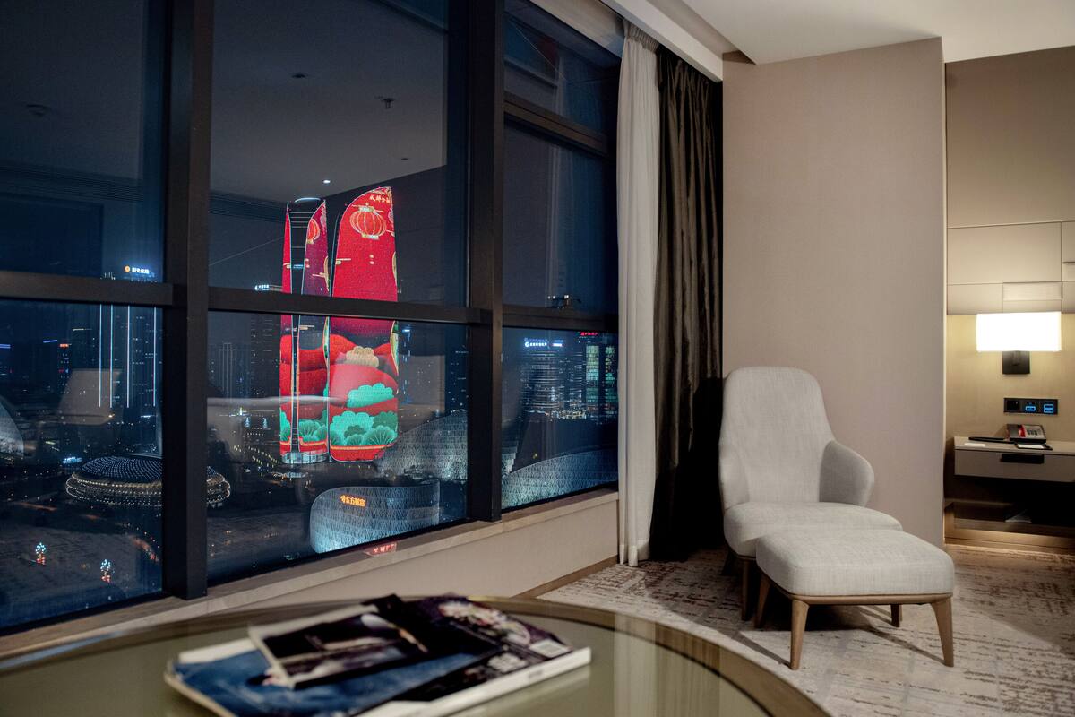Ein Hotelzimmer mit großem Fenster, einem weißen Sessel, einem kleinen Tisch mit einer Zeitschrift und einer Lampe auf dem Nachttisch.
