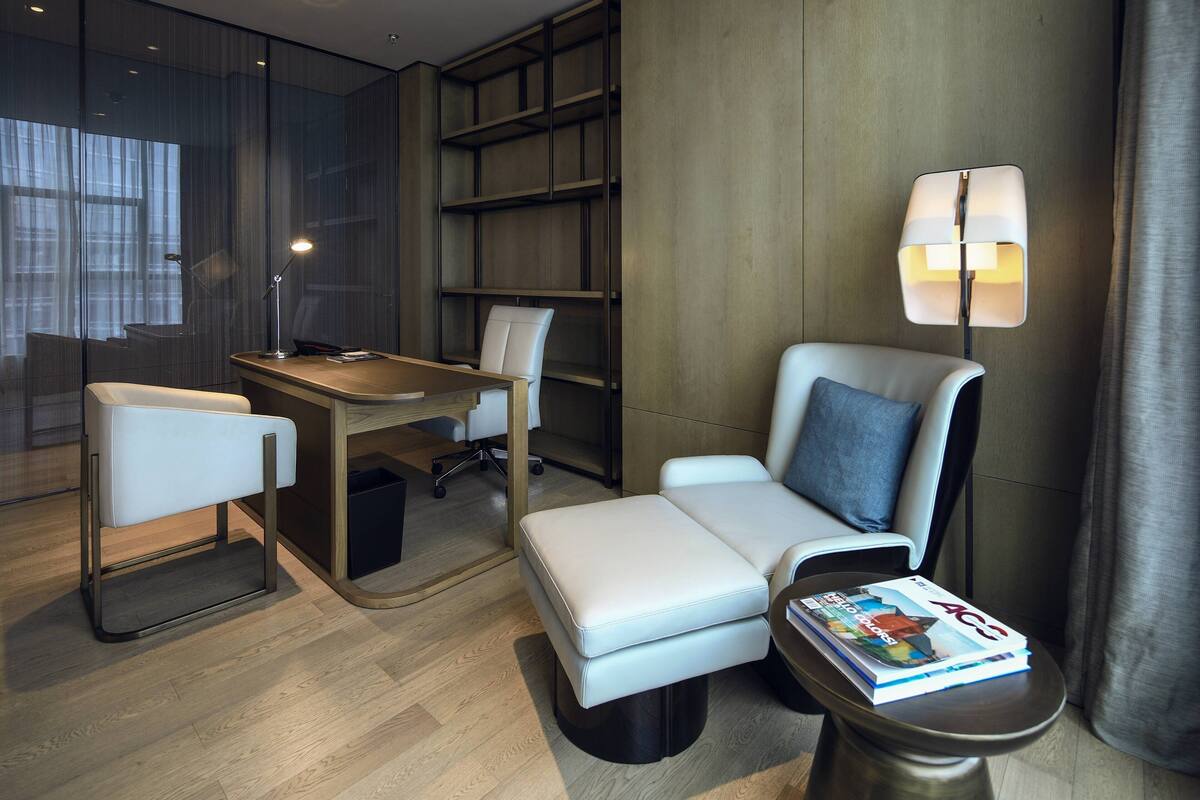 Ein modernes Hotelzimmer mit Schreibtisch, zwei Sessel, einer Couch, einem Bücherregal und einer Lampe.
