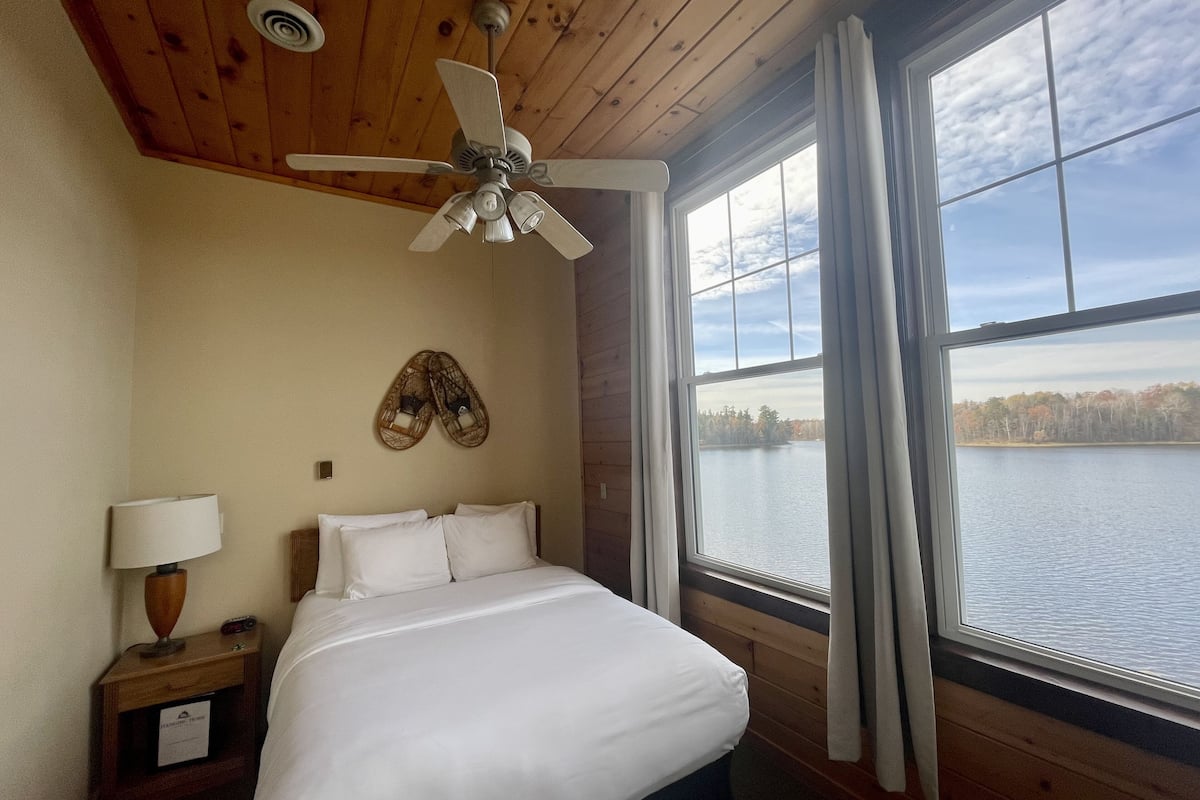 A bedroom with a bed, a ceiling fan, a nightstand with a lamp, a window with curtains, and a view of a lake.