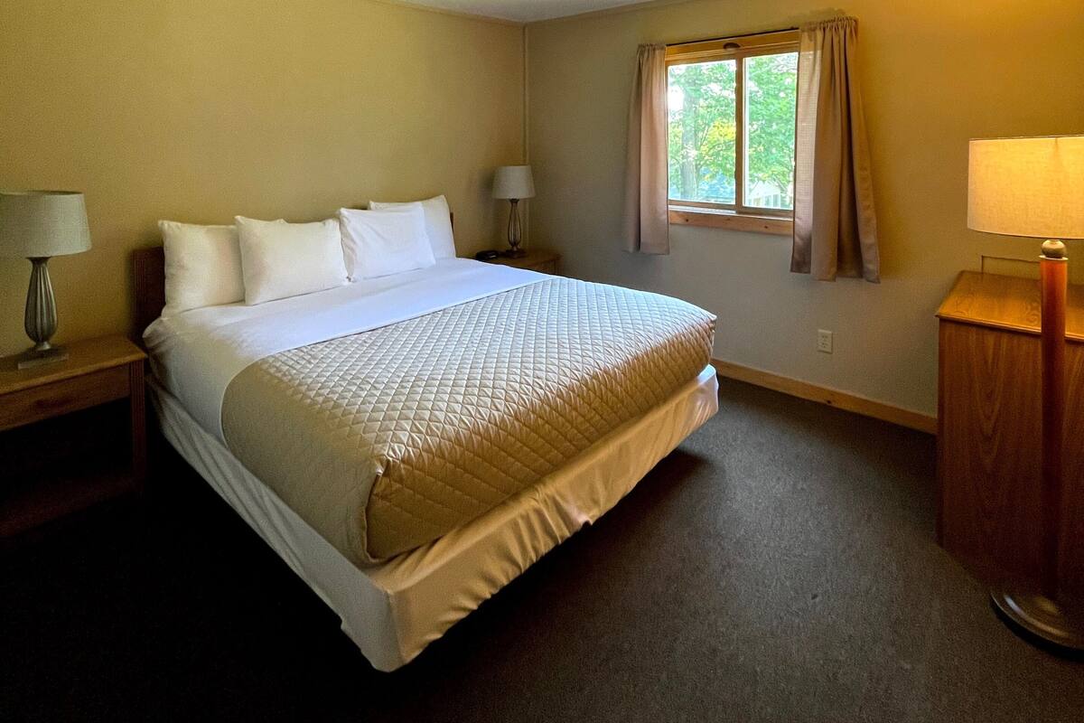 A neatly made bed in a hotel room with a window, a nightstand with a lamp, and a wooden side table.