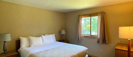 A neatly made bed in a hotel room with a window, a nightstand with a lamp, and a wooden side table.