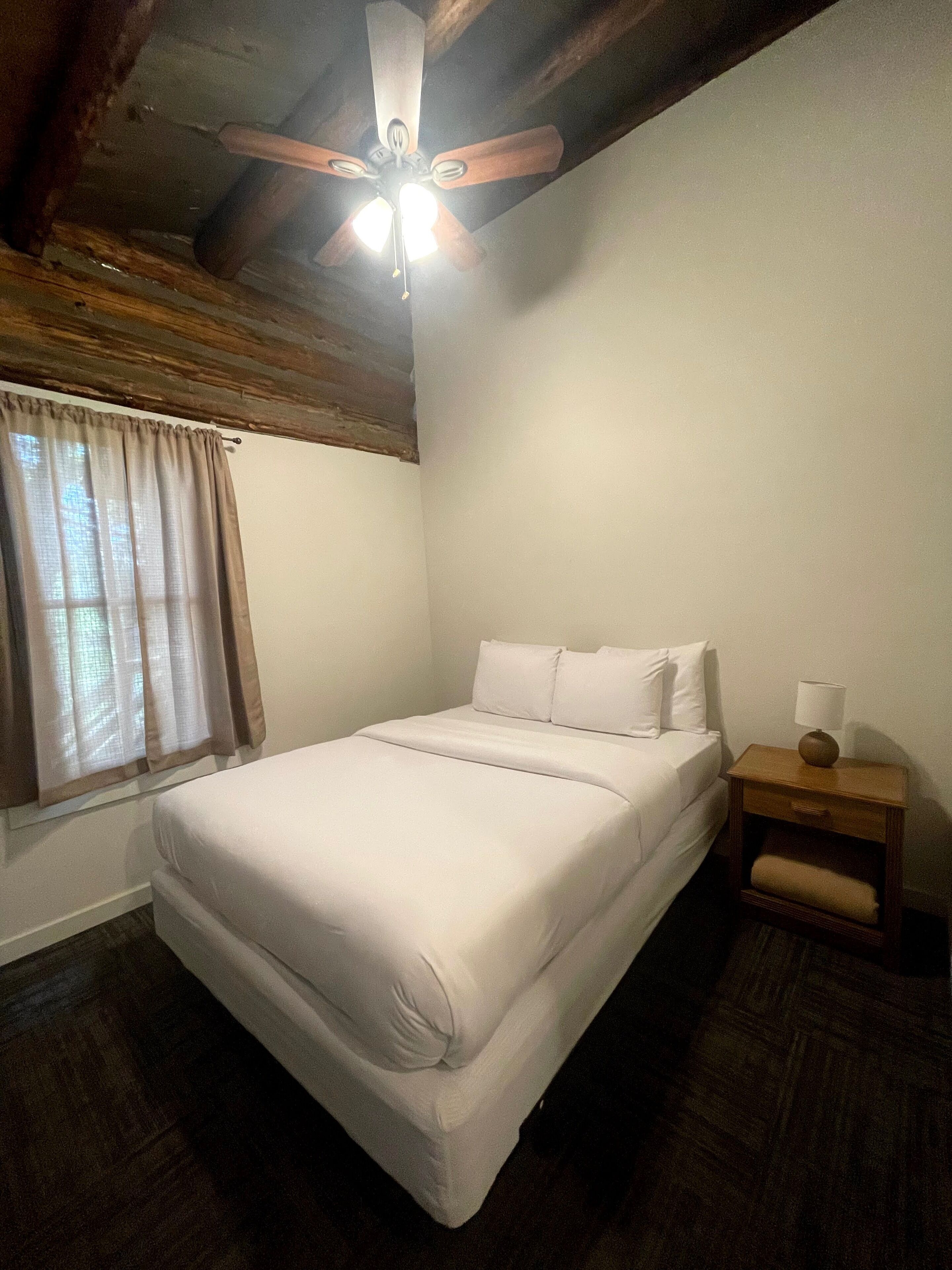 Une chambre à coucher avec un lit, une table de chevet, un ventilateur de plafond et une fenêtre avec des rideaux.