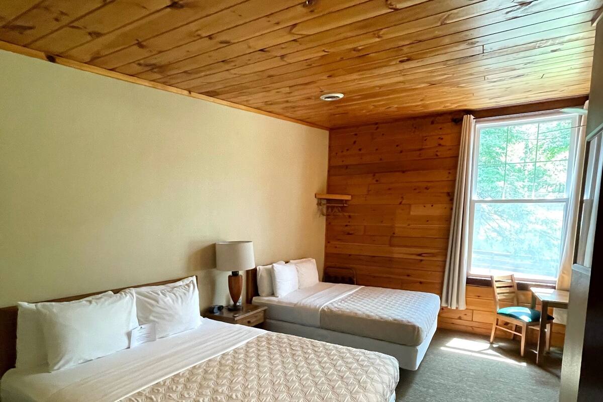 A room with two beds, a small table, a chair, and a window with a view of trees.