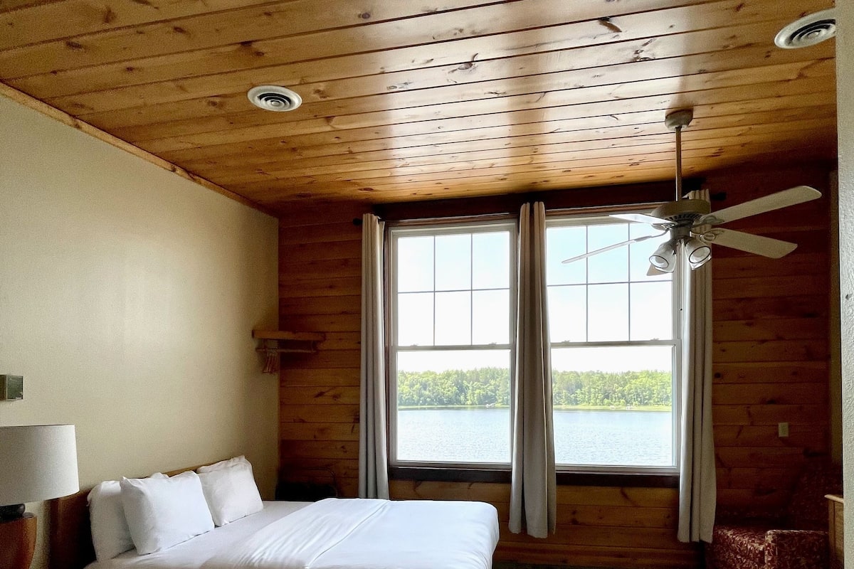 A room with two beds, a ceiling fan, a window with curtains, and a view of a body of water.
