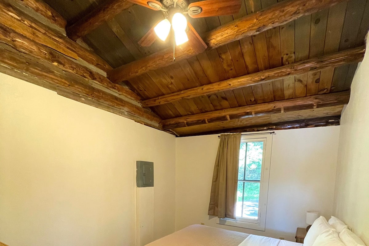 A bedroom with a wooden ceiling, a bed with white bedding, a window with curtains, and a small bedside table.