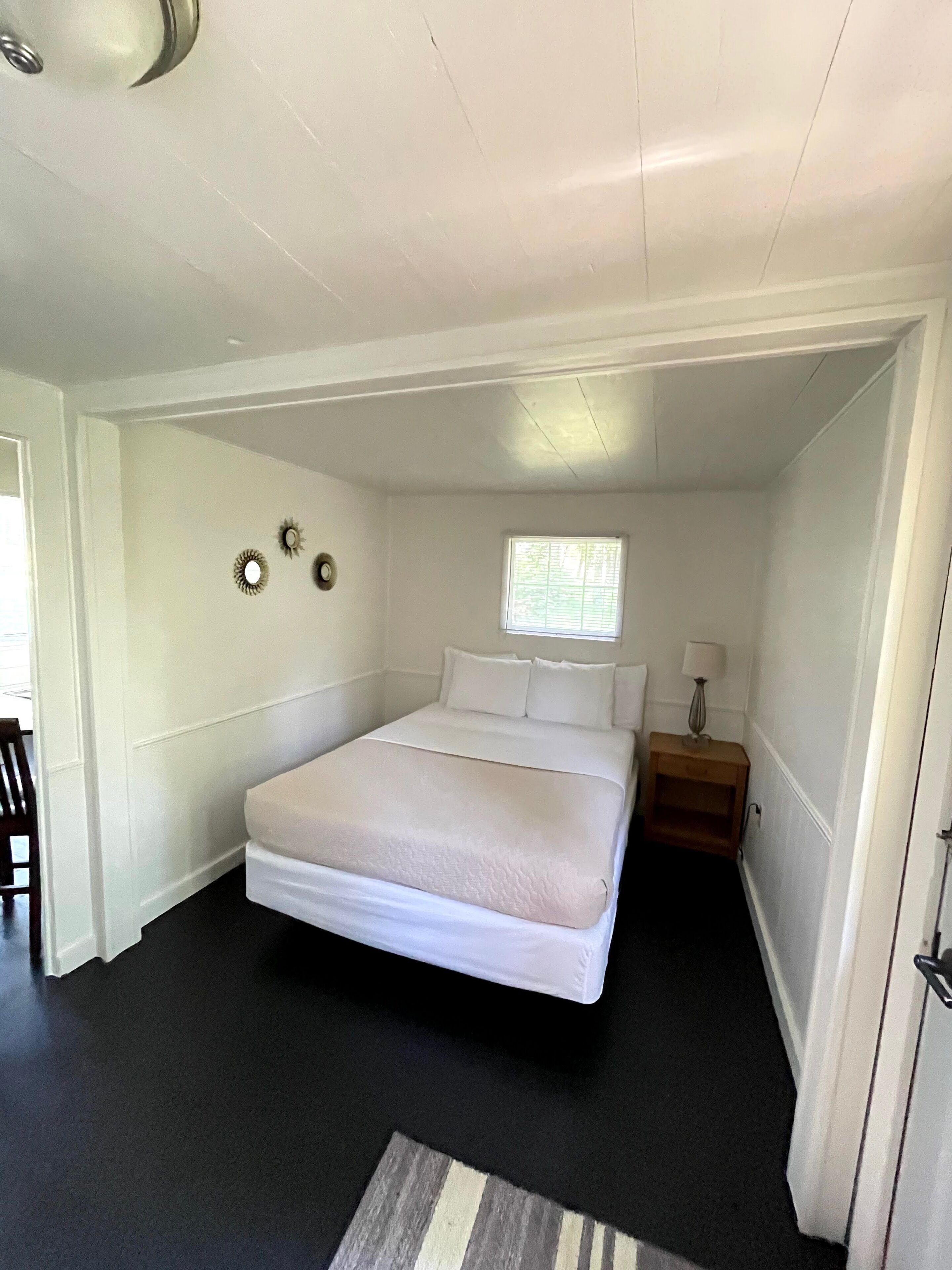 A bedroom with a large bed, a small bedside table with a lamp, a window, and a door leading to another room.