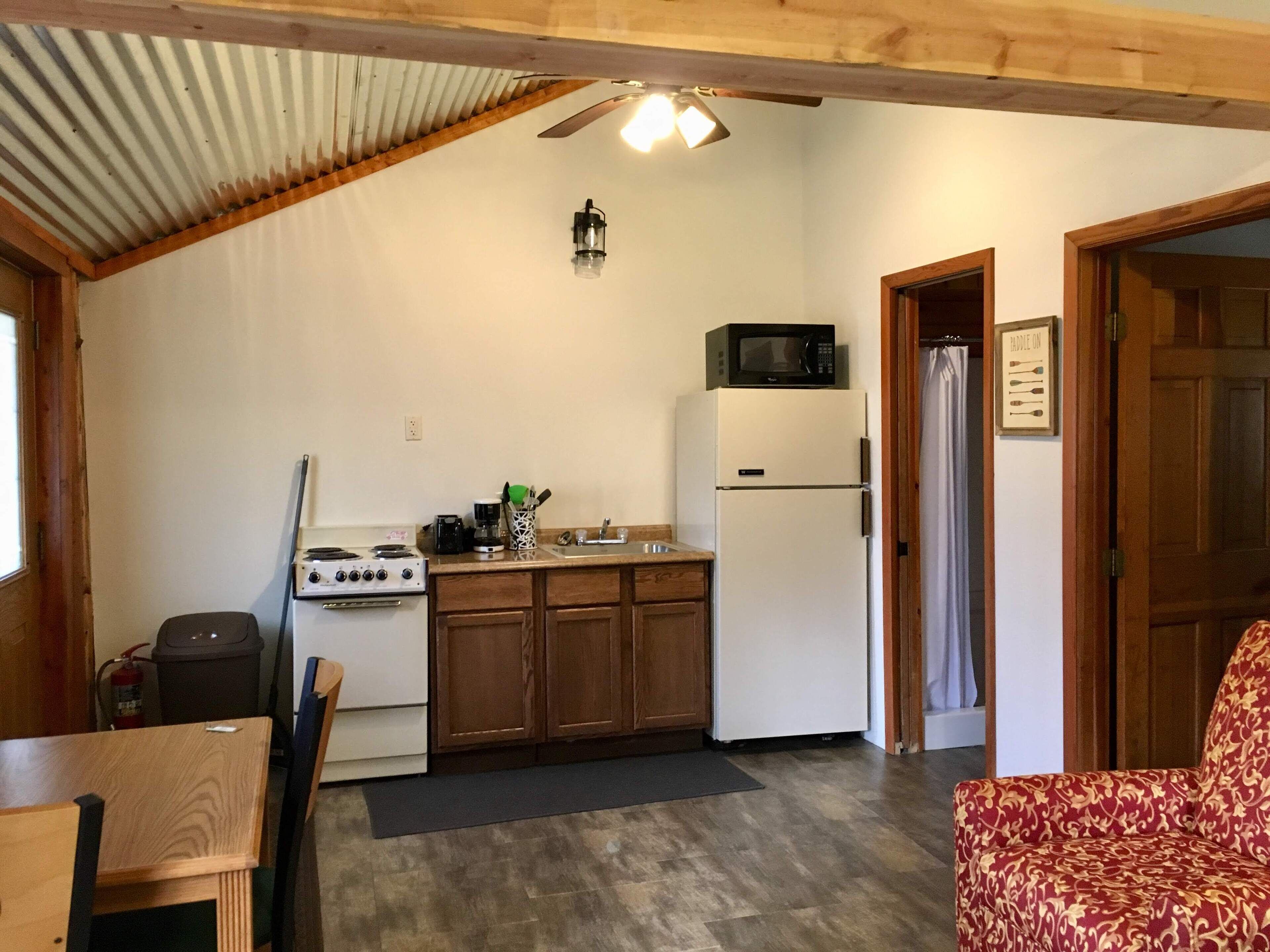 Cocina compacta con estufa, microondas y refrigerador, gabinetes de madera y un ventilador de techo.