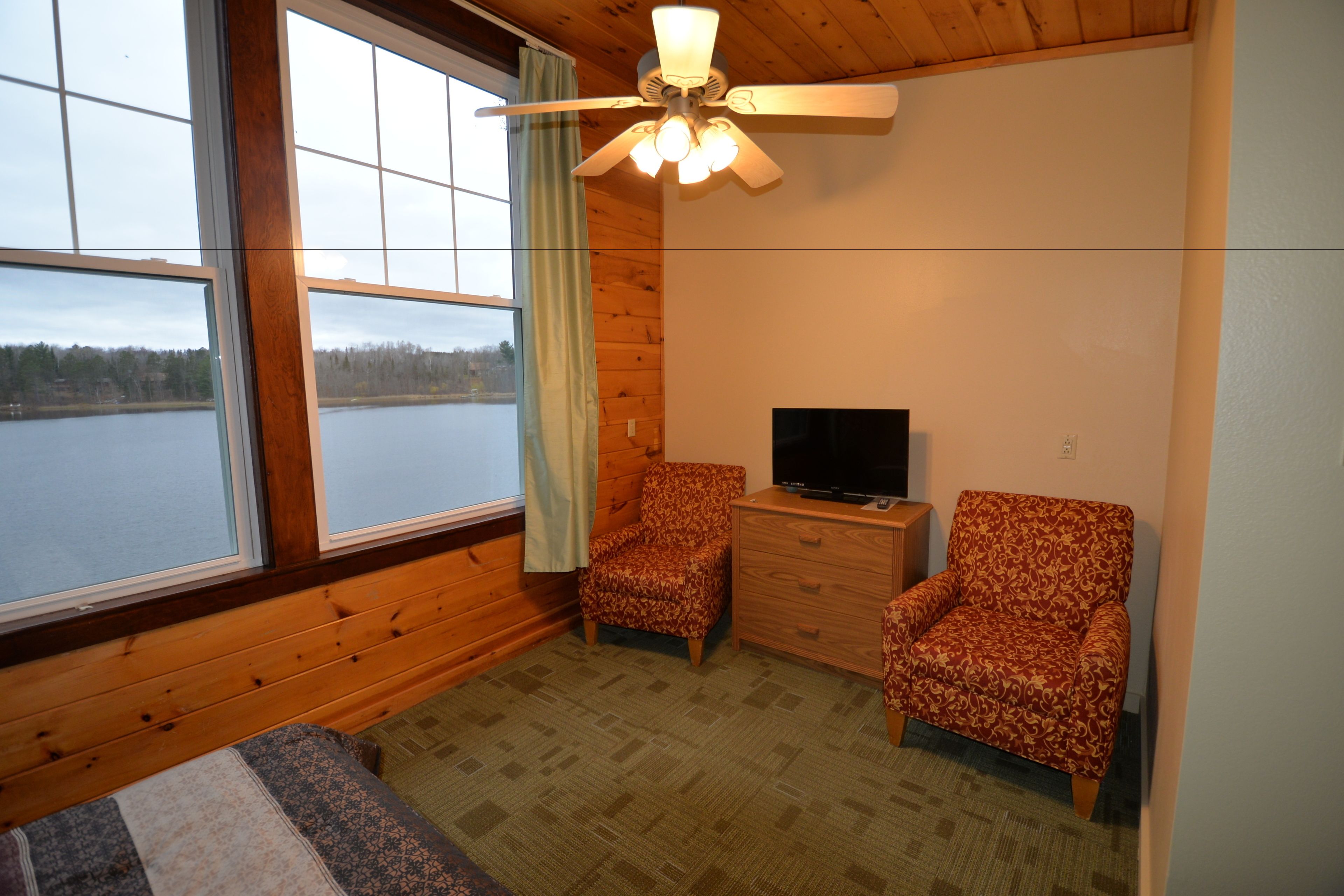 A room with a large window, a ceiling fan, two patterned armchairs, a TV on a wooden dresser, and a bed with a patterned bedspread.