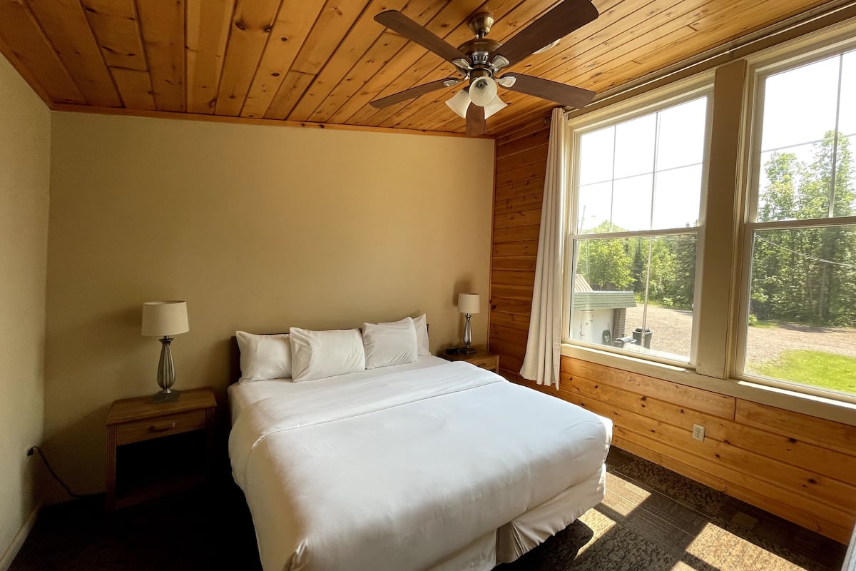 A bedroom with a bed, two bedside lamps, a ceiling fan, and a large window with a view of greenery.