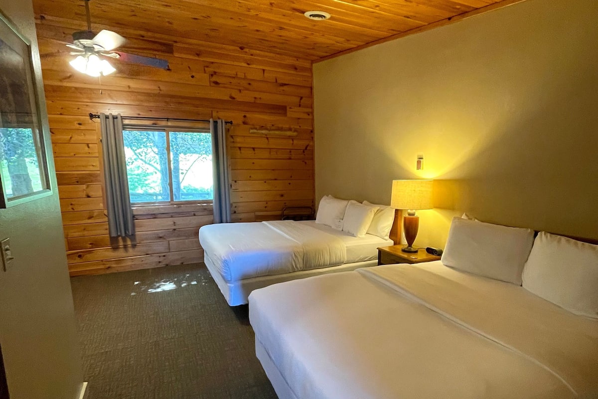 A room with two beds, a ceiling fan, a window with curtains, and a lamp on the nightstand.