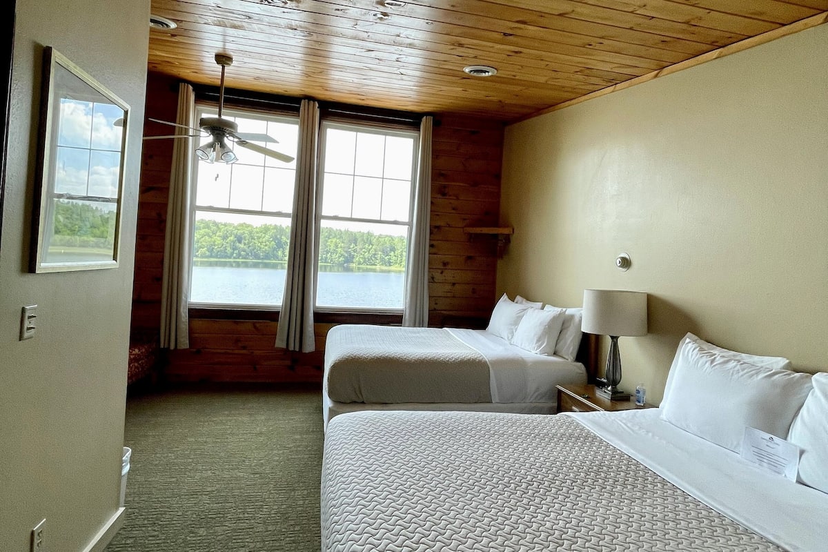 A hotel room with two beds, a ceiling fan, a window with curtains, and a view of the outdoors.