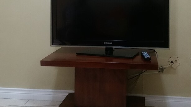 A flat-screen TV on a wooden stand in a room with tiled flooring. - Oasis at Oaklands (Kingston)