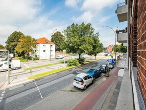 Vista de la calle con coches aparcados, un edificio blanco con techo rojo y una acera bordeada de árboles.