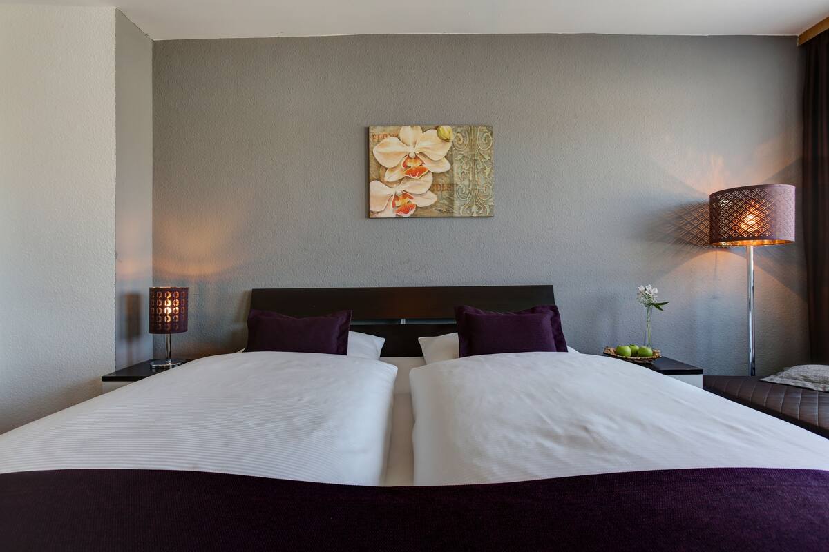 A neatly made bed with white linens and two purple pillows, a wall-mounted artwork of flowers, a bedside table with a lamp, and a small vase with flowers.