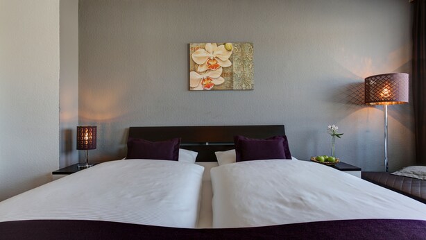 Una cama bien hecha con sábanas blancas y dos almohadas moradas, una obra de arte en la pared con flores, una mesita de noche con una lámpara y un pequeño jarrón con flores.