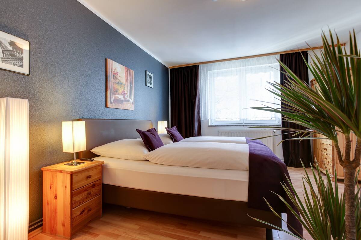 A bedroom with a bed, nightstand, lamp, and a large window with curtains.